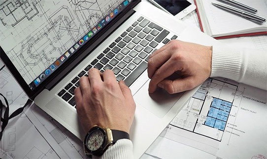 Hands typing on a laptop next to technical drawings and plans on a desk